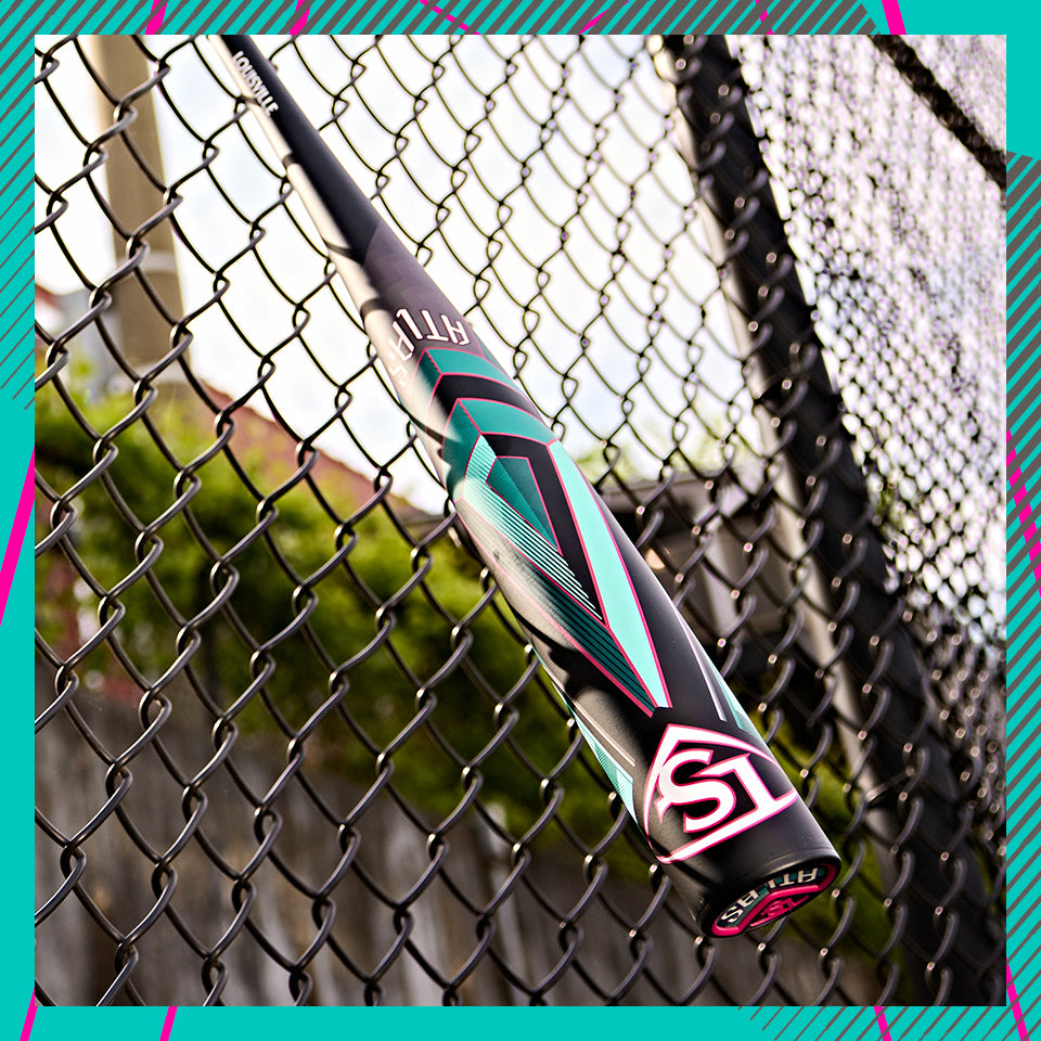 A close-up of a black baseball bat with teal, pink, and white geometric designs leaning against a black chain-link fence, with greenery and concrete visible in the background.