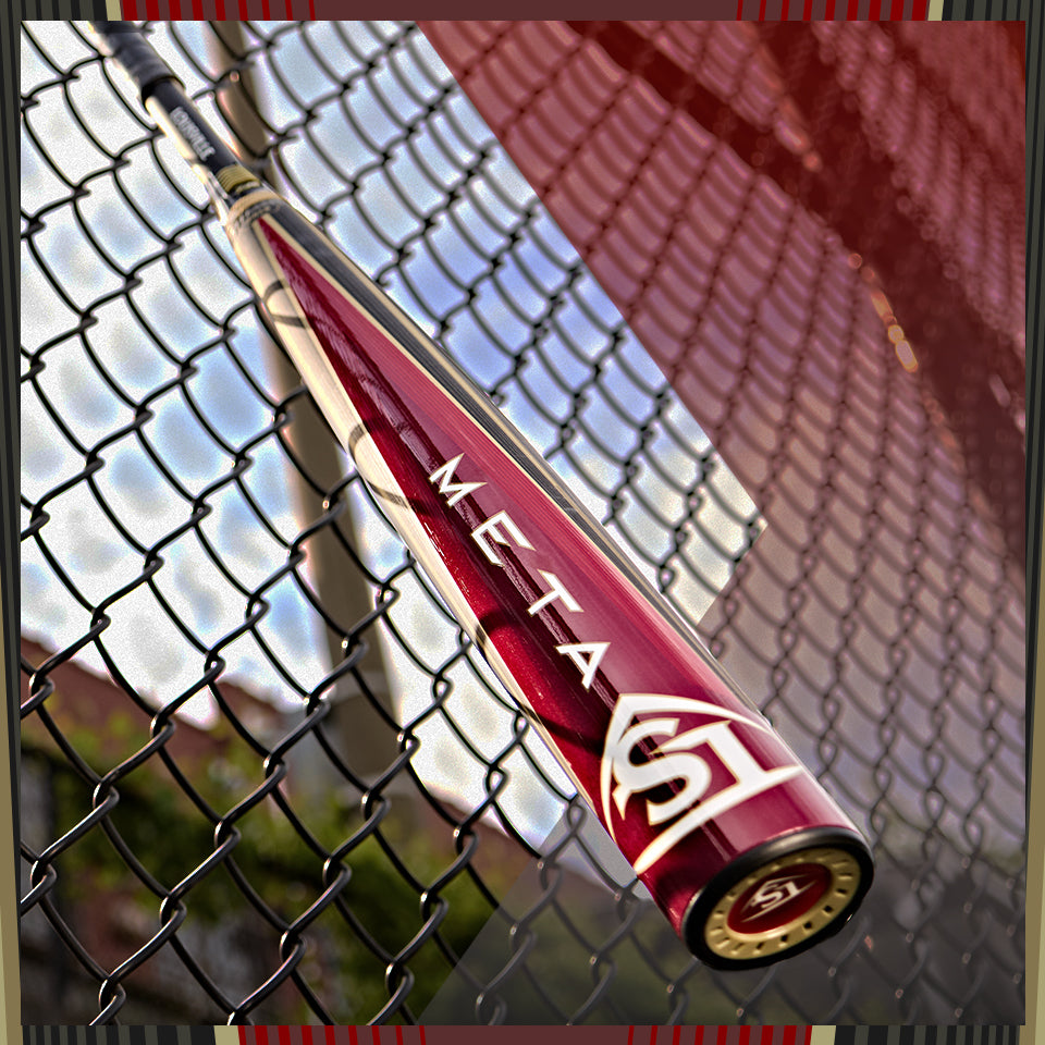 A close-up of a maroon and silver Meta SL baseball bat resting against a chain-link fence, with a blurred background of a baseball field and sky.