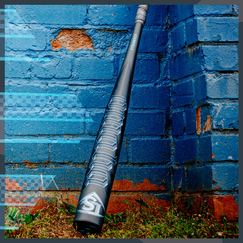 A black and silver baseball bat labeled “Louisville Slugger” leans against a weathered blue brick wall with patches of exposed red brick and grass at the base.