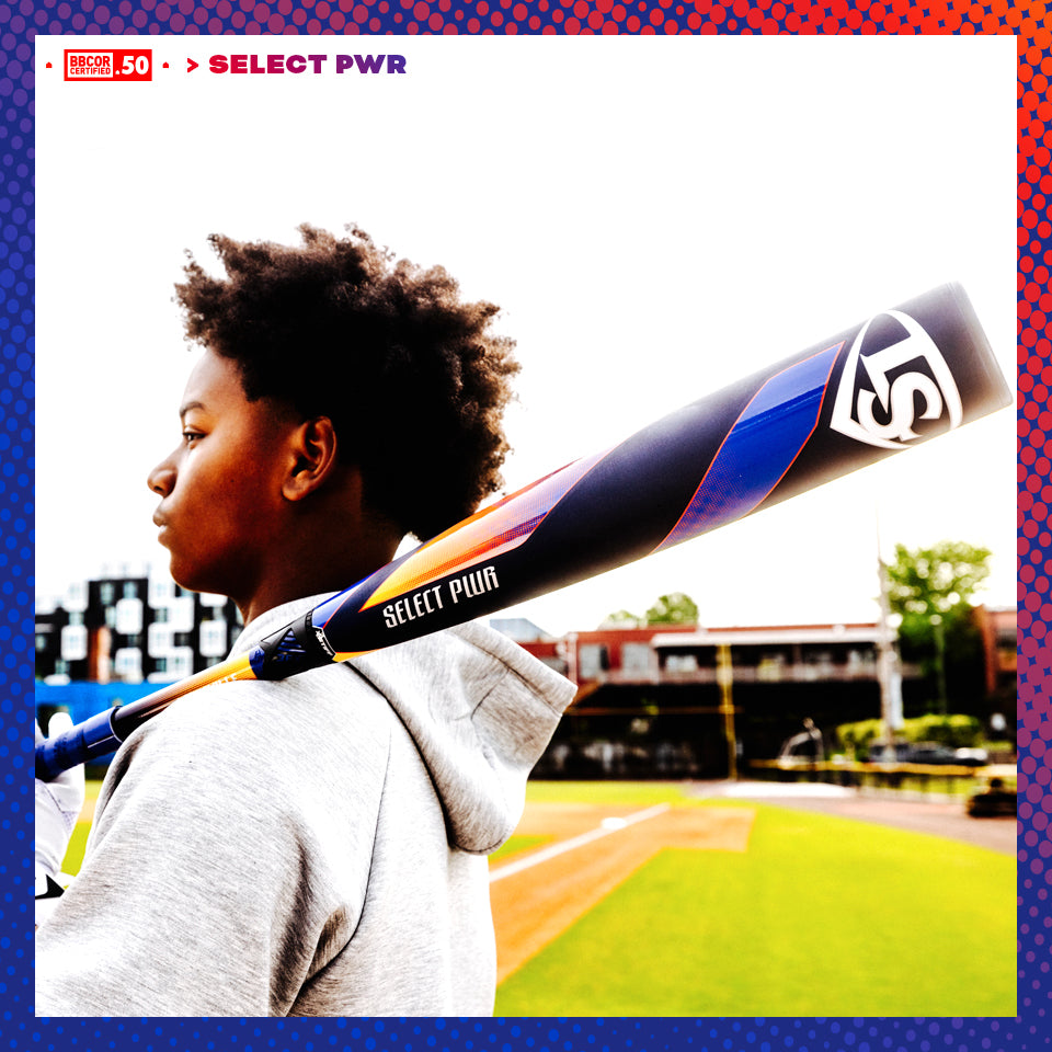 A young baseball player stands on a field with the 2025 Louisville Slugger Select PWR (-3) BBCOR Baseball Bat (WBL2967010 DEMO) resting on his shoulder, gazing into the distance with stadium seating and buildings behind him under a bright sky.