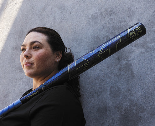 A woman with dark hair holds the 2025 Louisville Slugger LXT (-10) Fastpitch Softball Bat: WBL2994010 (DEMO) over her shoulder, standing before a gray textured wall and gazing thoughtfully into the distance.
