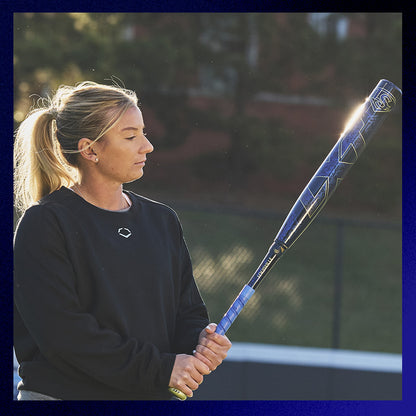 A woman with blonde hair in a ponytail, wearing a black sweatshirt, stands outdoors holding a Louisville Slugger 2025 LXT (-9) Fastpitch Softball Bat (WBL2995010), looking down as sunlight shines behind her.
