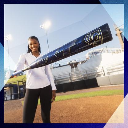 A smiling woman in a white shirt and black pants stands on a baseball field, holding the Louisville Slugger 2025 LXT (-11) Fastpitch Softball Bat (WBL2993010) toward the camera, with empty bleachers and stadium lights behind her.