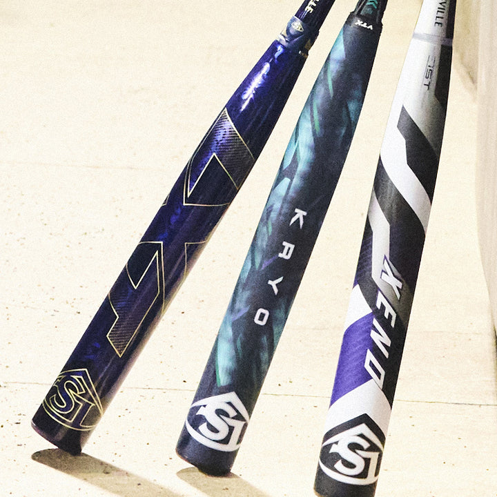 Three colorful baseball bats with distinct designs and brand logos are standing upright on a light-colored surface, leaning against a wall.