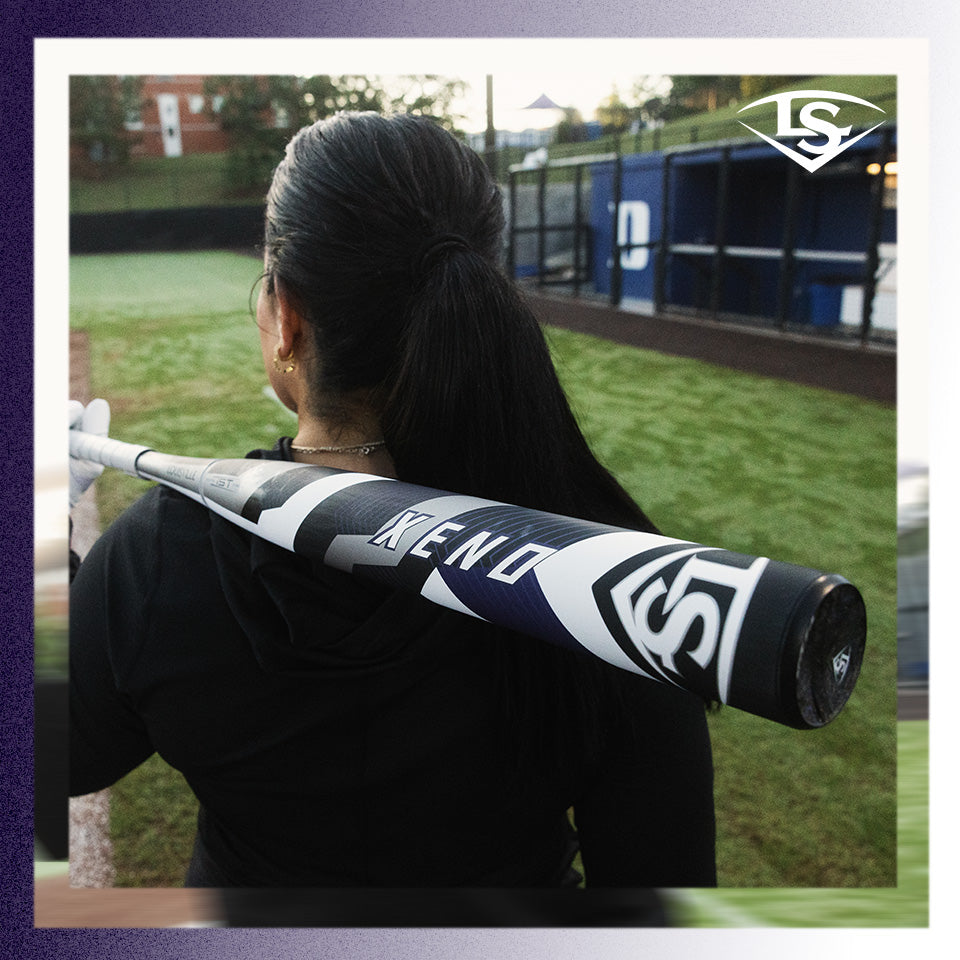 A person with long dark hair in a ponytail stands on a baseball field, holding a black and white Louisville Slugger bat labeled XENO over their shoulder. The field and dugout are visible in the background.