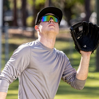 A baseball player in a gray shirt, black cap, and Franklin MLB Pro Sunglasses (24012C) looks up while preparing to catch a ball with a black glove on a sunny day outdoors.