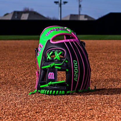 A Wilson A2000 1786SS 11.5" Baseball Glove (WBW103218115), with black, green, and pink accents, stands upright on infield dirt, with blurred houses and a fence in the background.