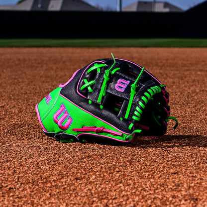 A Wilson A2000 1786SS 11.5" Baseball Glove (WBW103218115) rests on infield dirt, with a blurred background of a fence and houses. This Summer 2025 Wilson glove features black, green, and pink accents.