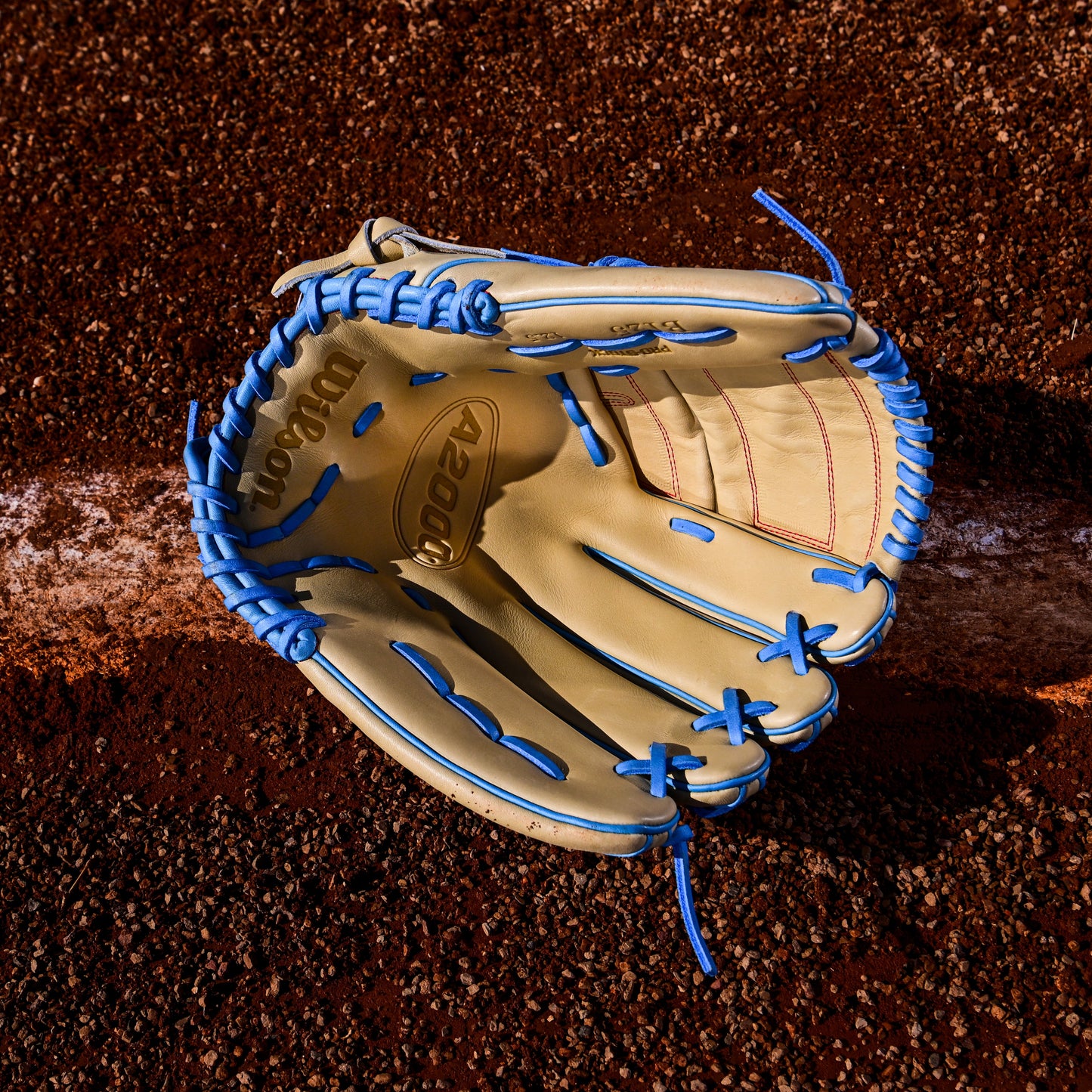 A tan Wilson A2000 B125 12.5" Baseball Glove (WBW103220125) with blue laces lies open on a brown dirt field near a white chalk baseline. Crafted from Pro Stock leather, this Wilson glove ensures premium durability and performance.