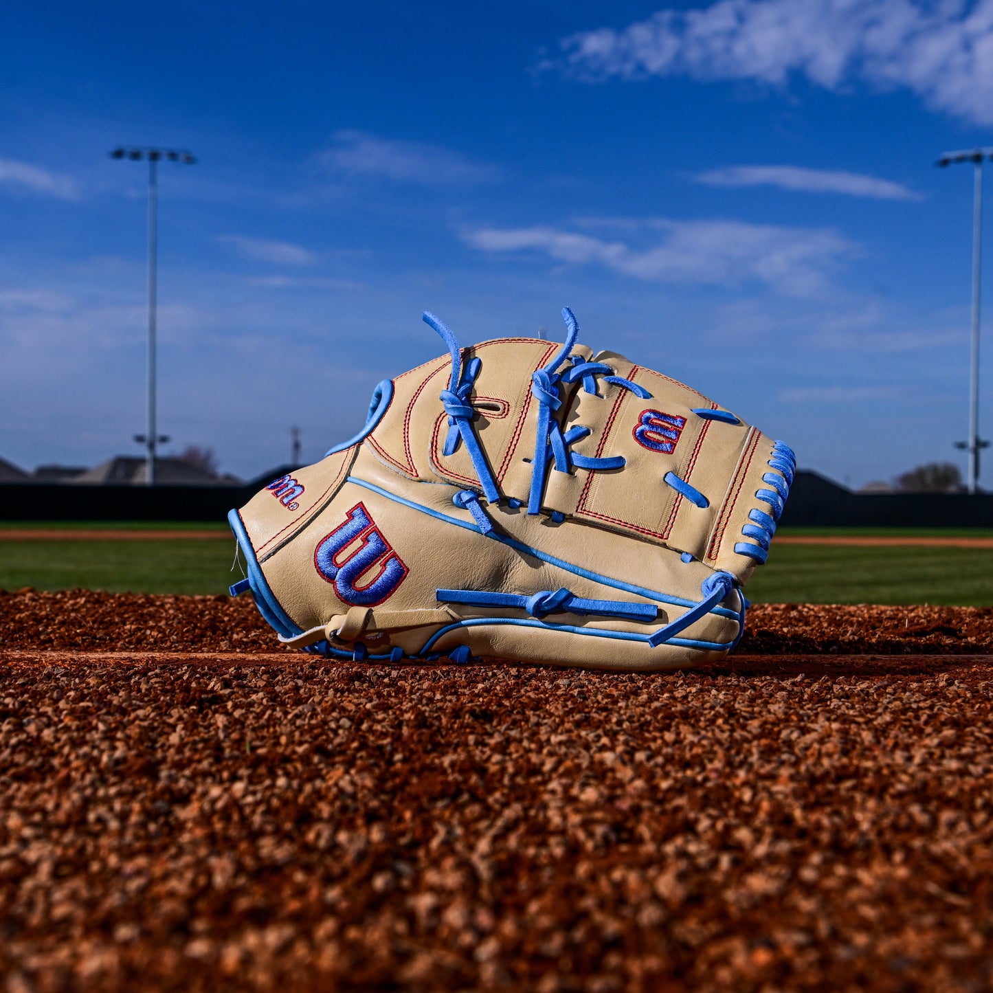 A Wilson A2000 B125 12.5" baseball glove (WBW103220125) with blue laces and red accents rests on the field's dirt beneath a bright blue, cloud-streaked sky.