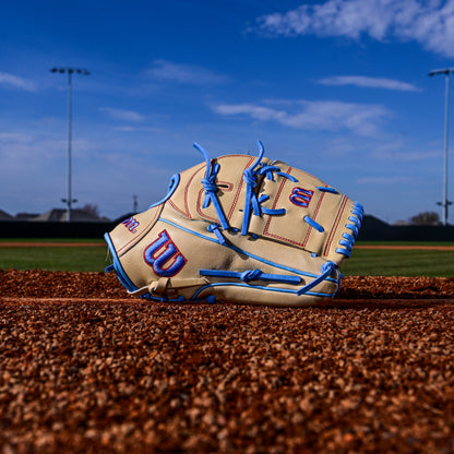 A Wilson A2000 B125 12.5" baseball glove (WBW103220125) with blue laces and red accents rests on the field's dirt beneath a bright blue, cloud-streaked sky.