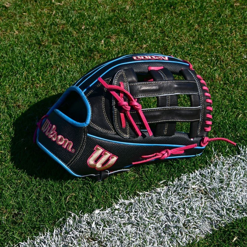 A Wilson A2000 1777SS 12.75" Baseball Glove (WBW1032221275), black with SuperSkin, blue trim, and pink laces, rests on green grass near a white chalk line on a sports field.