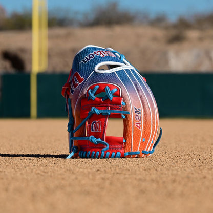 The Wilson A2K 1786SS 11.5" Baseball Glove (WBW103290115) features vibrant red, blue, and orange accents and stands upright on the infield dirt, with blurred green fencing and foul poles in the background.
