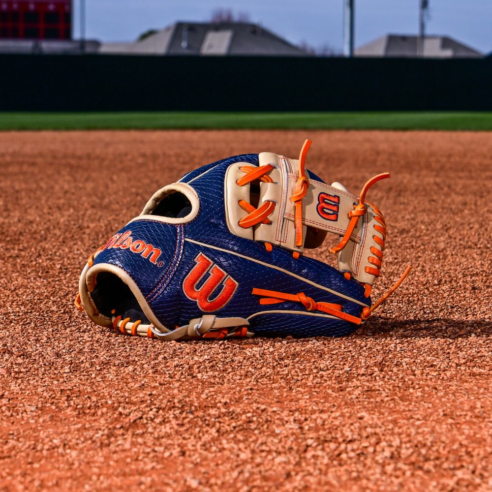 A Wilson A2000 JA27 11.5" Jose Altuve GM Baseball Glove (Summer 2025: WBW103295115) with blue, cream, and orange Pro Stock leather lies on the infield dirt, grass and buildings in the background.