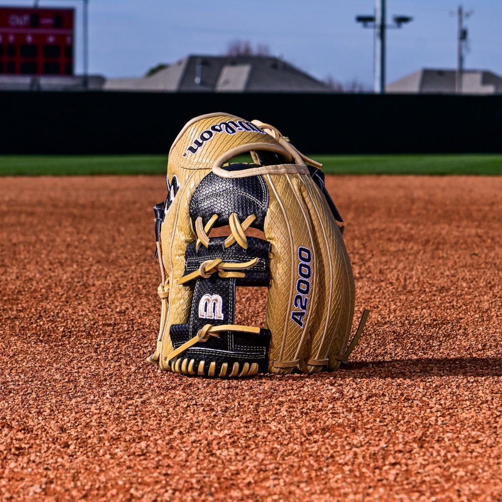 A Wilson A2000 KBH13 11.75" Ke'Bryan Hayes GM Baseball Glove (WBW1032961175) stands upright on infield dirt, with grass, a scoreboard, and buildings blurred in the background under a blue sky.