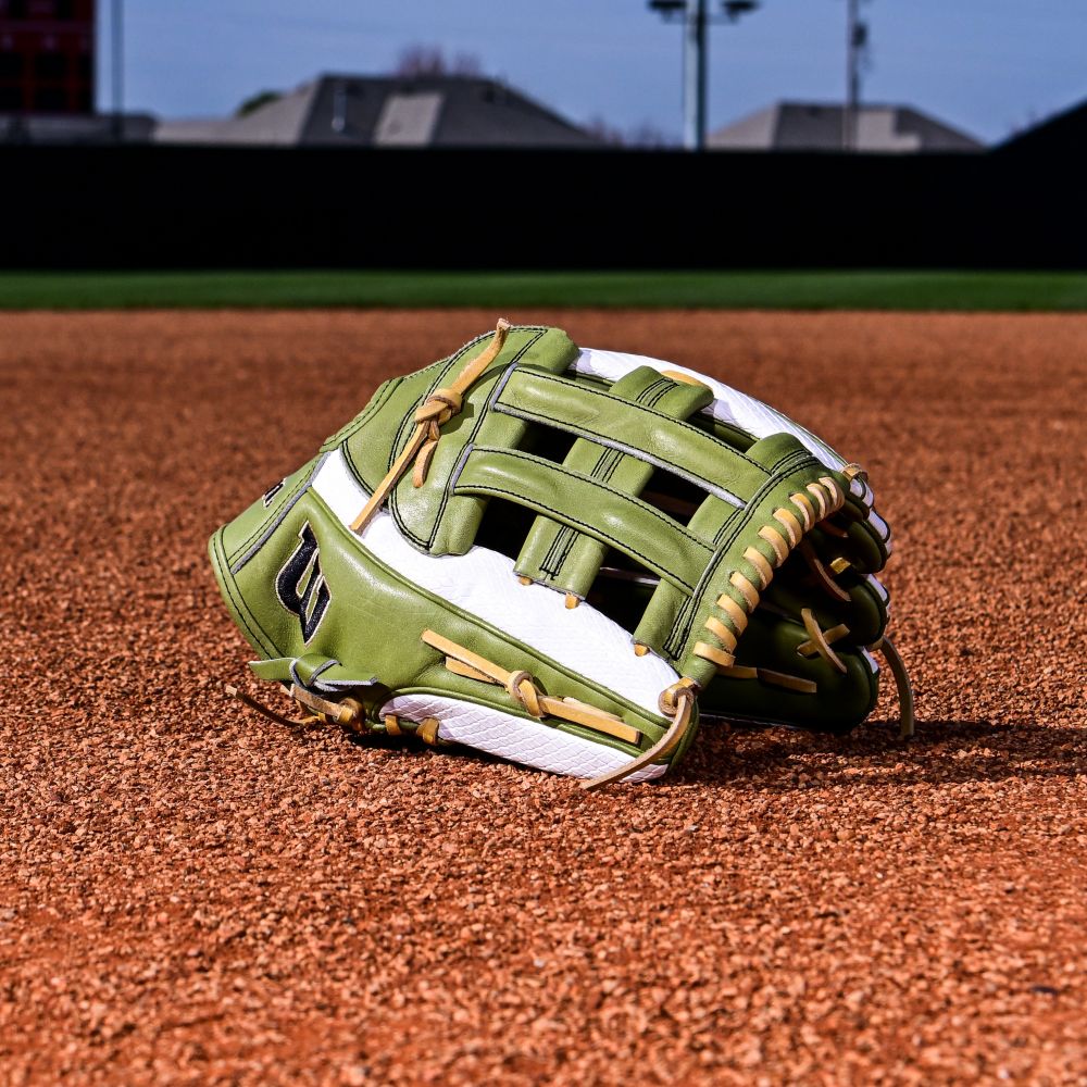 A Wilson A2000 FP50SS 12.25" Fastpitch Glove (Summer 2025) in green and white with a Dual Post Web lies on reddish-brown infield dirt, with grass and buildings visible in the background.