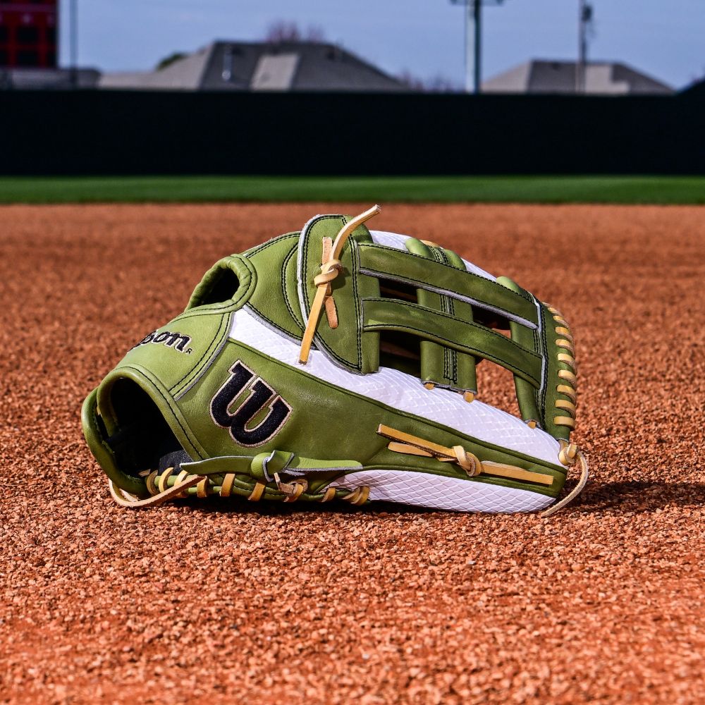 A Wilson A2000 FP50SS 12.25" Fastpitch Glove (green/white with tan laces and Dual Post Web) rests on infield dirt, framed by grass, buildings, and a blue sky.