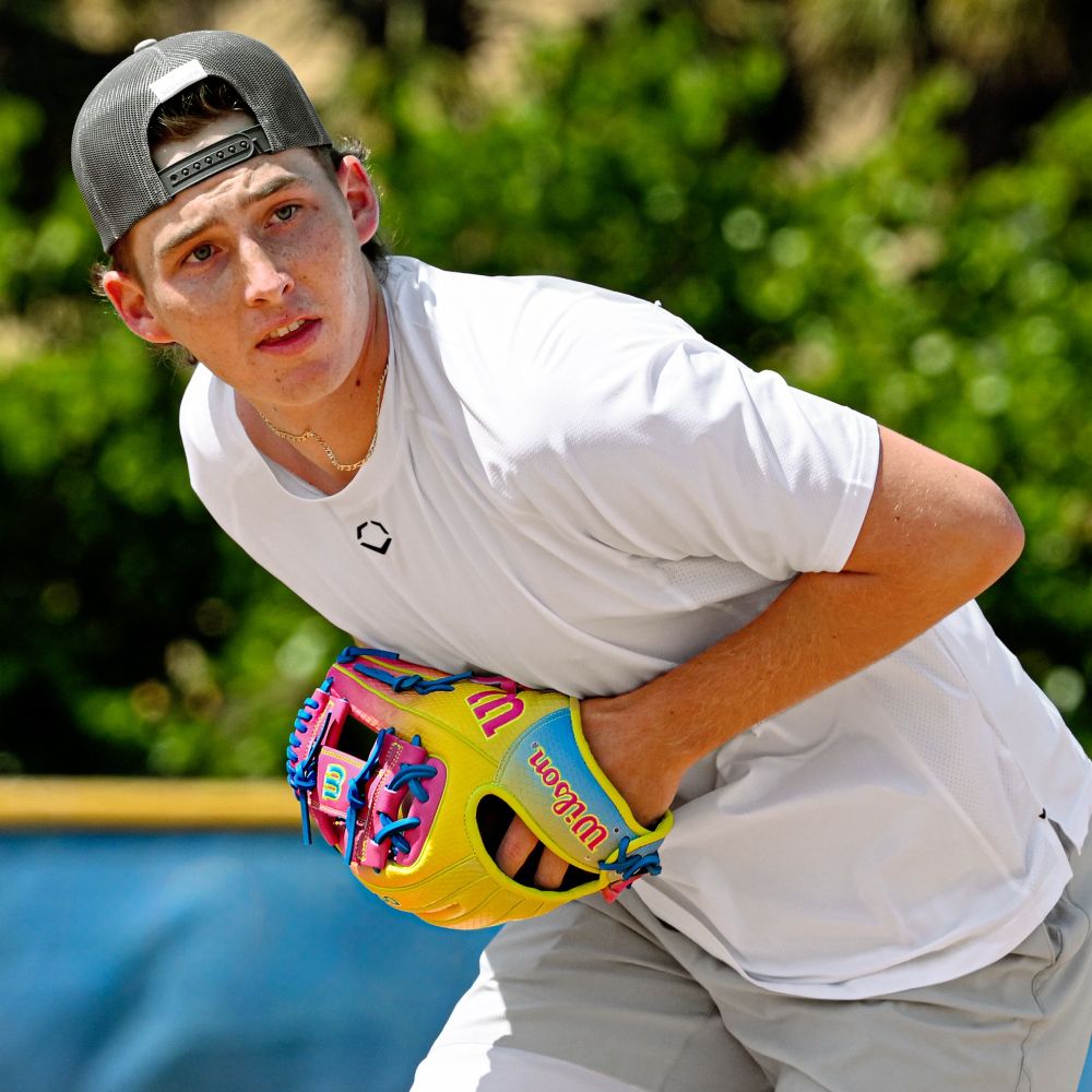 A baseball player in a white shirt, gray shorts, and backward cap readies to pitch, holding a Wilson A2000 1786SS 11.5" Baseball Glove (WBW103492115). Green foliage and a blue fence are visible in the background.