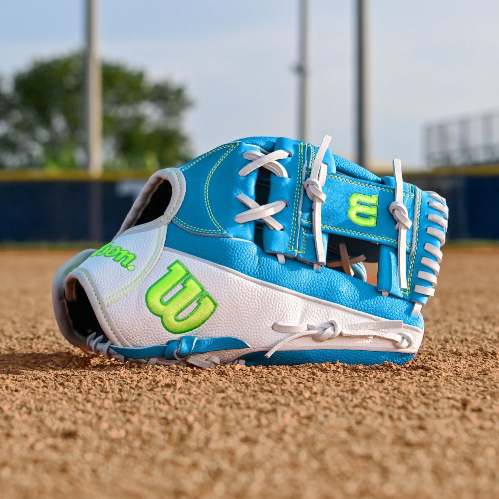A Wilson A2000 H12SS 12" Fastpitch Glove (WBW10348612) rests on a dirt field, blue and white with green accents. Trees, a fence, and poles appear blurred in the background.