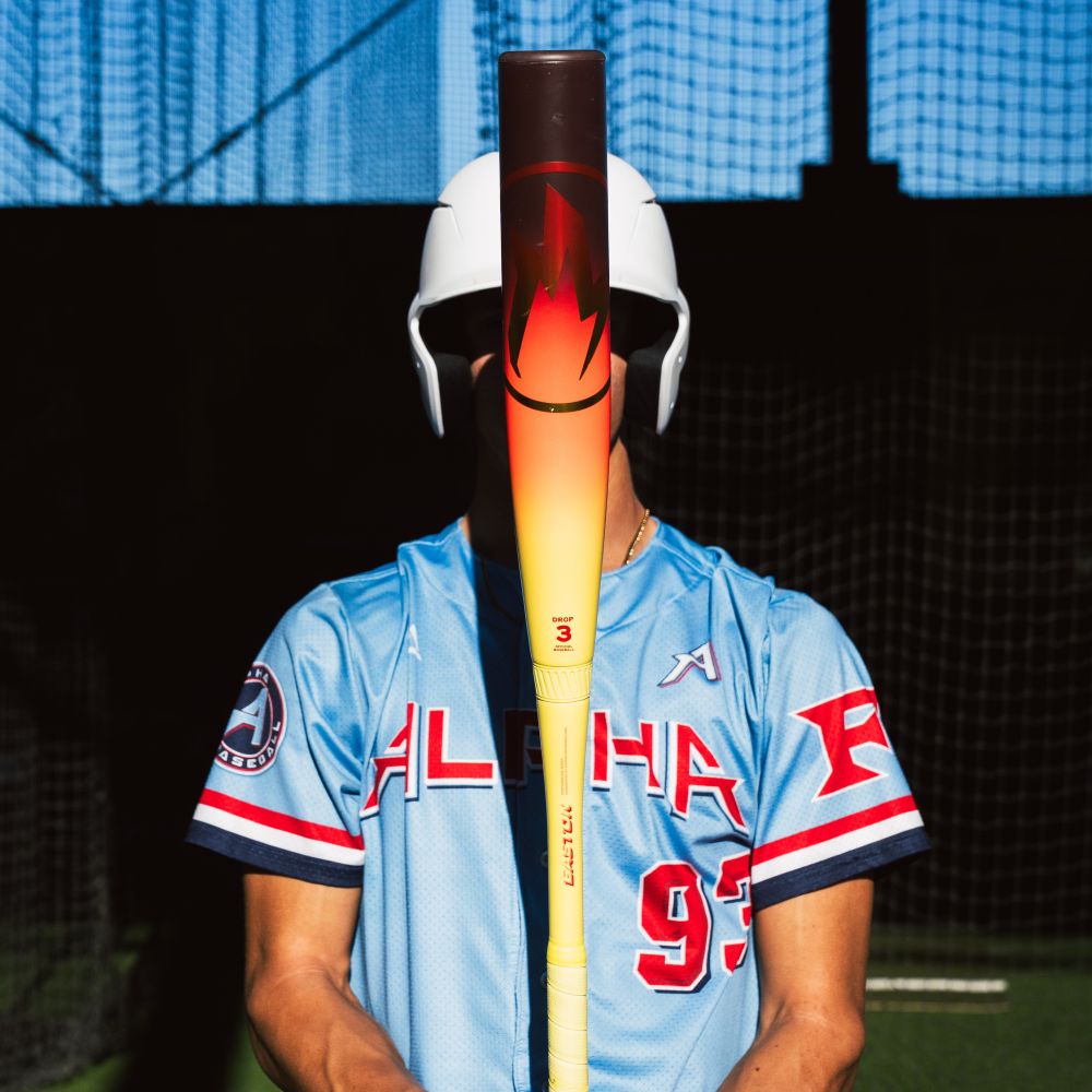 A baseball player in a light blue jersey and white helmet stands indoors, holding an Easton 2026 Hype Fire (-3) BBCOR Baseball Bat (EBB6HYP3) with a composite barrel vertically in front of their face, obscuring their features.