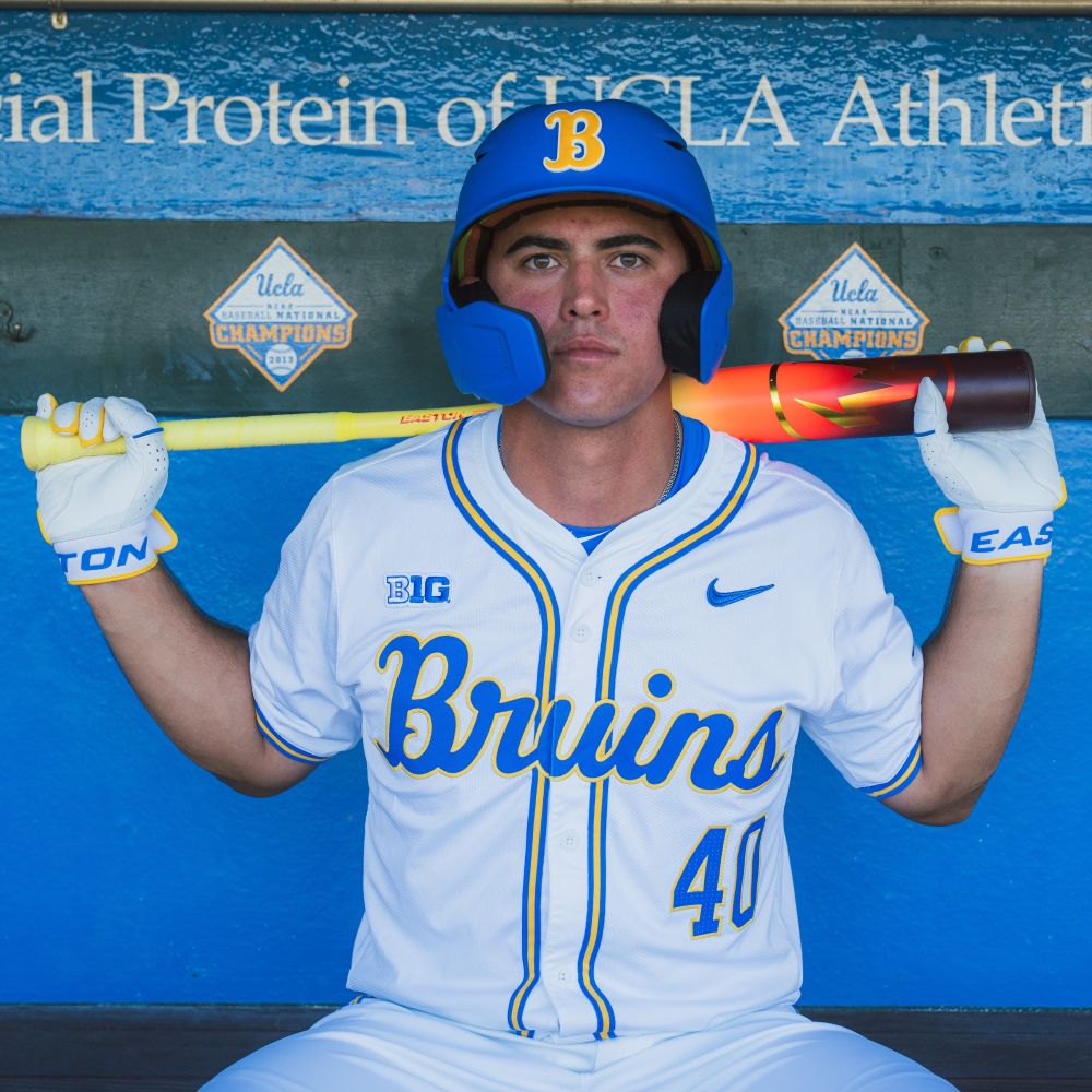 A UCLA Bruins baseball player in a white uniform and blue helmet sits in the dugout, holding the 2026 Easton Hype Fire (-3) BBCOR Baseball Bat (EBB6HYP3) across his shoulders. UCLA signs and athletic achievements are visible in the background.