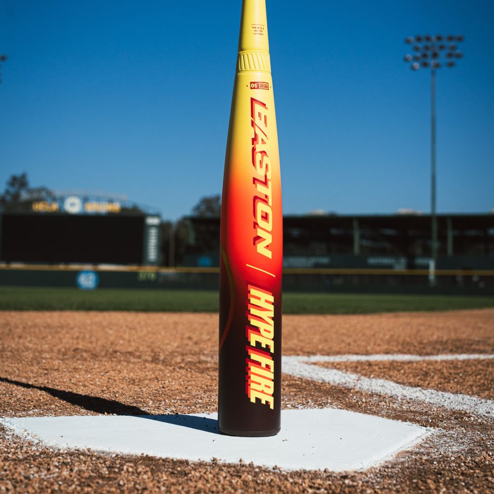 A yellow and red 2026 Easton Hype Fire (-3) BBCOR Baseball Bat (EBB6HYP3) by Easton stands upright on home plate, its composite barrel lit by stadium lights with a scoreboard in the background.