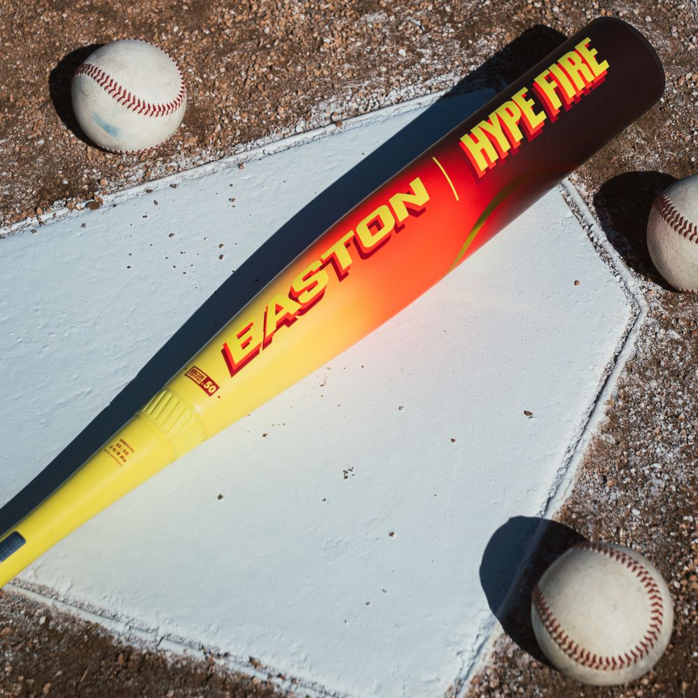 A yellow and red Easton 2026 Hype Fire (-3) BBCOR Baseball Bat (EBB6HYP3), with a composite barrel, lies across home plate beside four baseballs on the dirt infield.