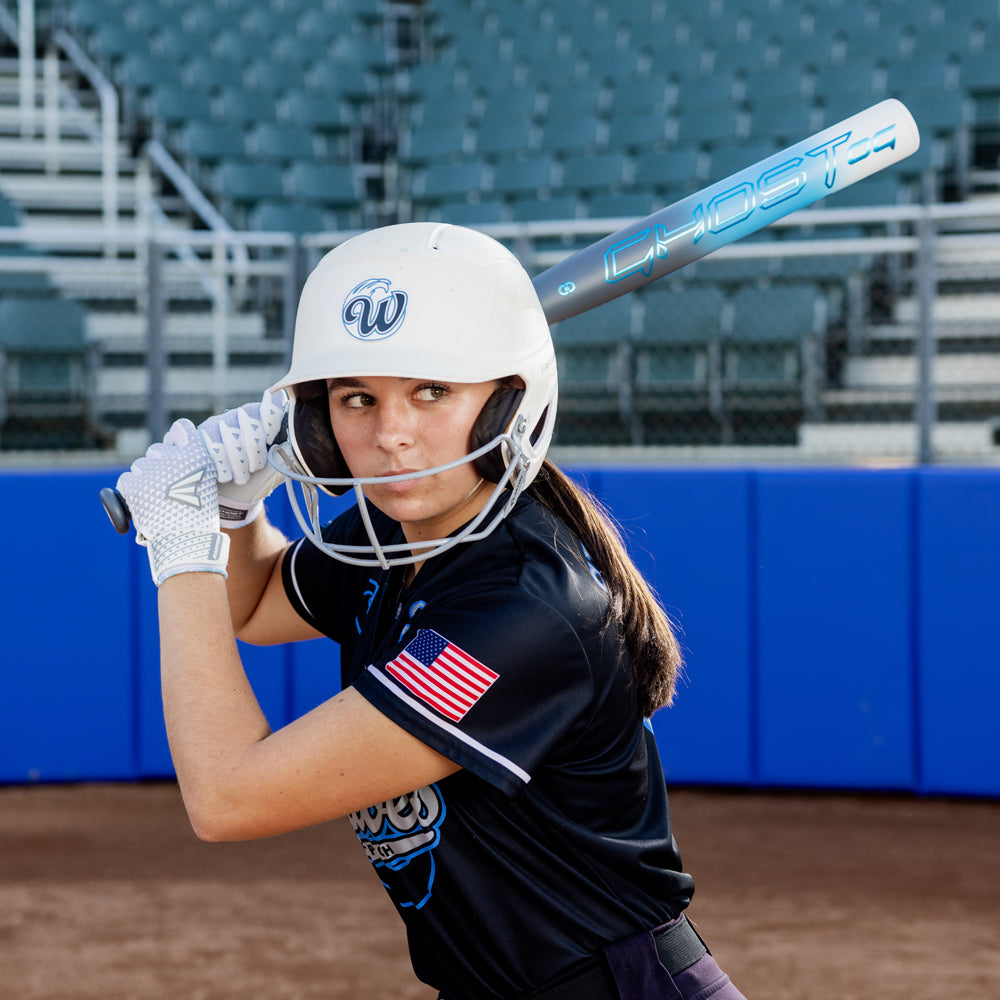 A softball player in a black uniform and white helmet gets ready to bat, holding the 2025 Easton Ghost OG (-11) Fastpitch Softball Bat (EFP5GHOG11), with empty stadium seats in the background.