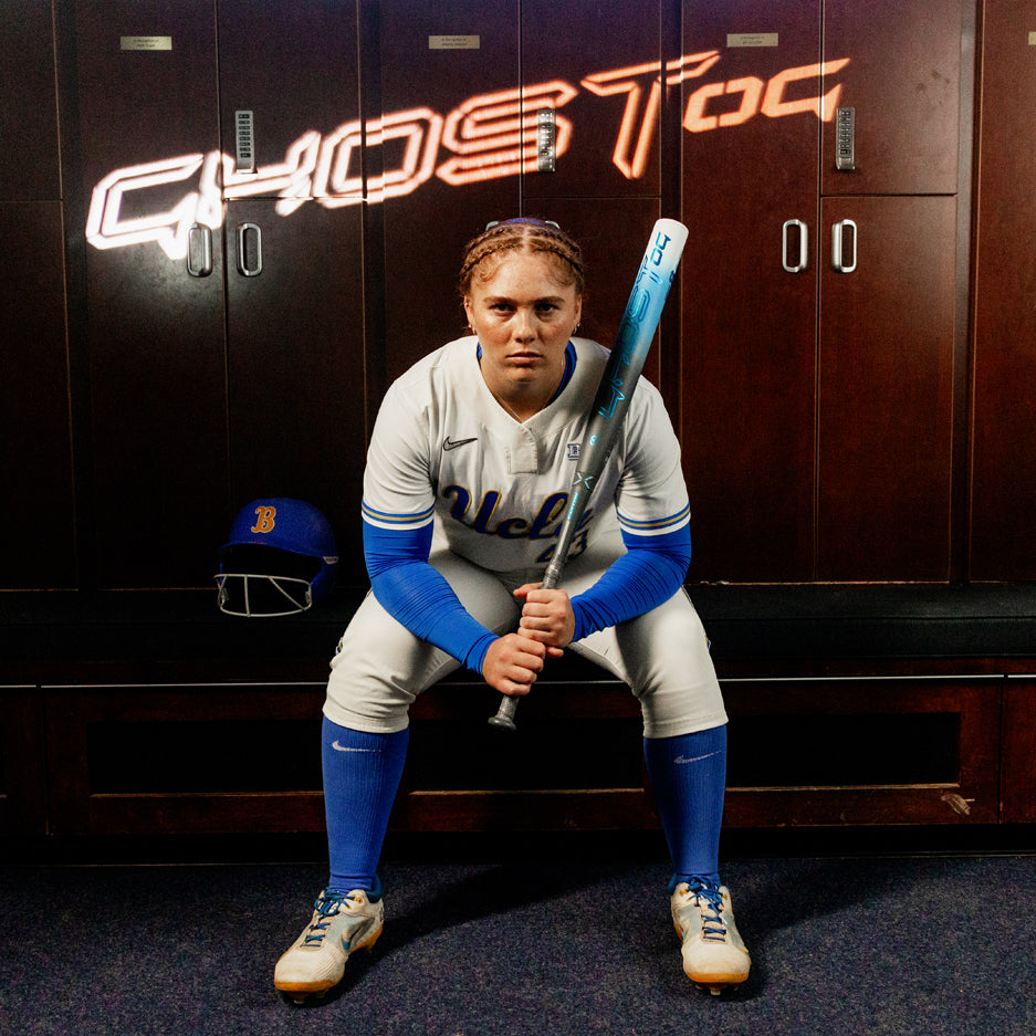 A UCLA softball player sits on a locker room bench, holding the 2025 Easton Ghost OG (-11) Fastpitch Softball Bat by Easton, with a helmet beside her and the glowing word "GHOST" above her head.