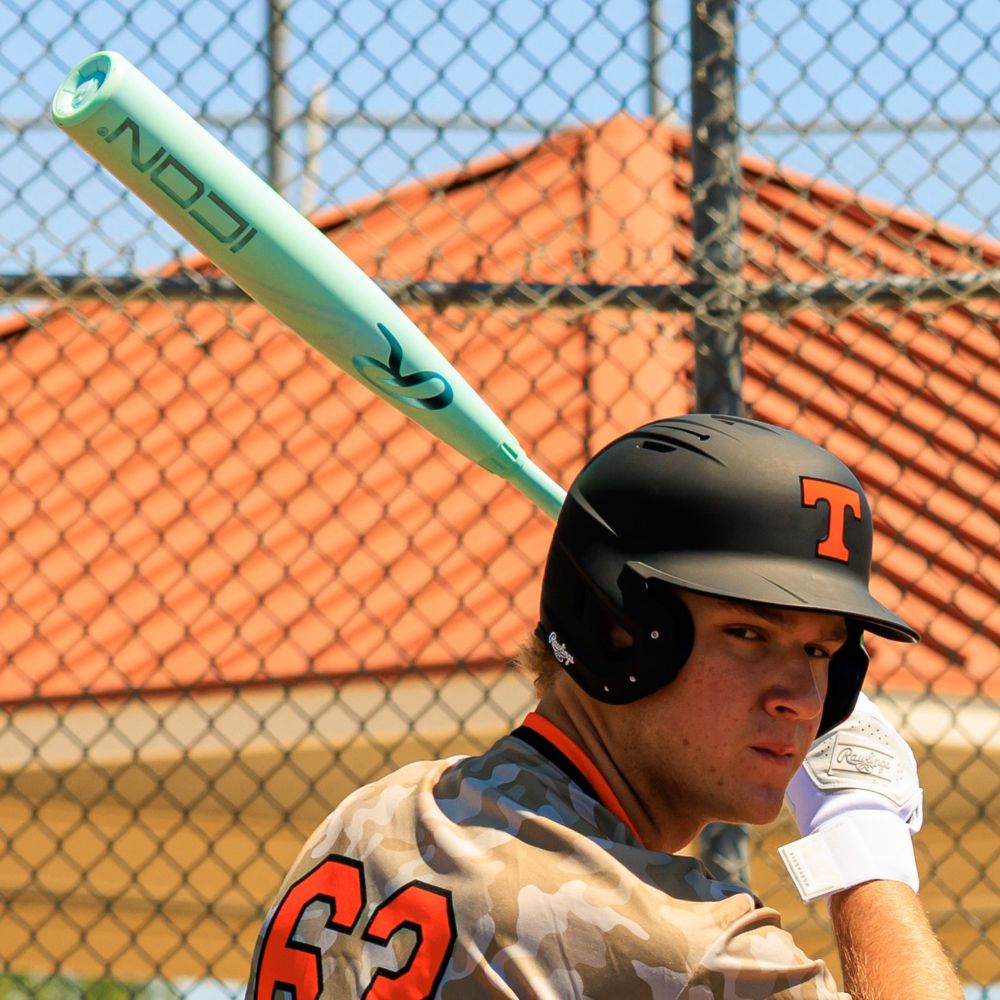 A baseball player in a black helmet with an orange T, camo jersey #62, and white gloves grips a mint green Rawlings 2026 Icon (-3) BBCOR Baseball Bat (RBB6I3). A chain-link fence and red-roofed building are visible behind him.