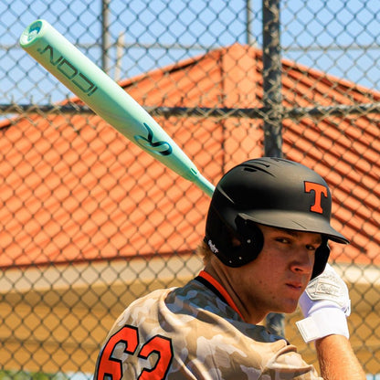 A baseball player in a black helmet with an orange T, camo jersey #62, and white gloves grips a mint green Rawlings 2026 Icon (-3) BBCOR Baseball Bat (RBB6I3). A chain-link fence and red-roofed building are visible behind him.