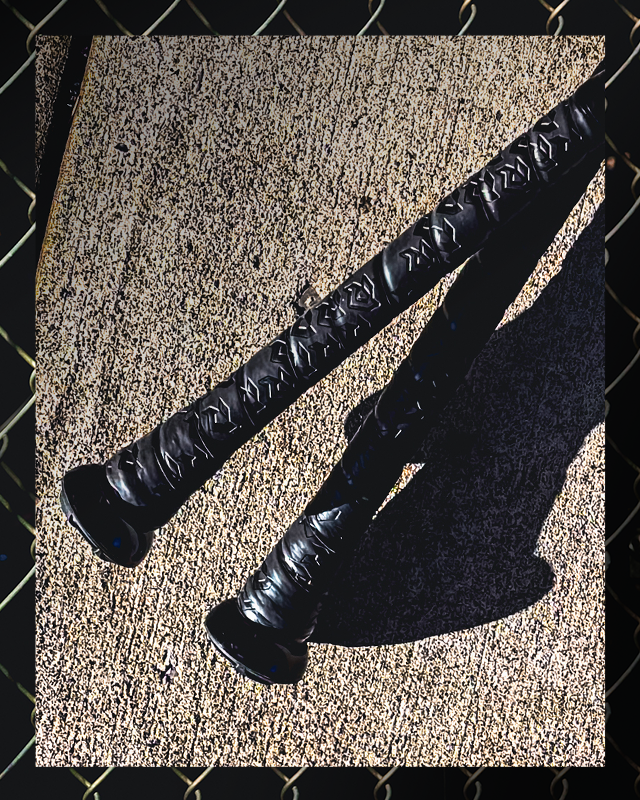 Two DeMarini 2026 The Goods (-3) BBCOR Baseball Bats (WBD2588010) lie parallel on concrete, casting shadows, with a blurred chain-link fence framing the foreground.