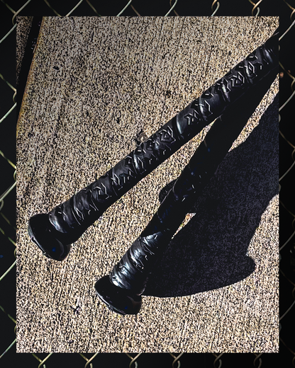 Two DeMarini 2026 The Goods (-3) BBCOR Baseball Bats (WBD2588010) lie parallel on concrete, casting shadows, with a blurred chain-link fence framing the foreground.