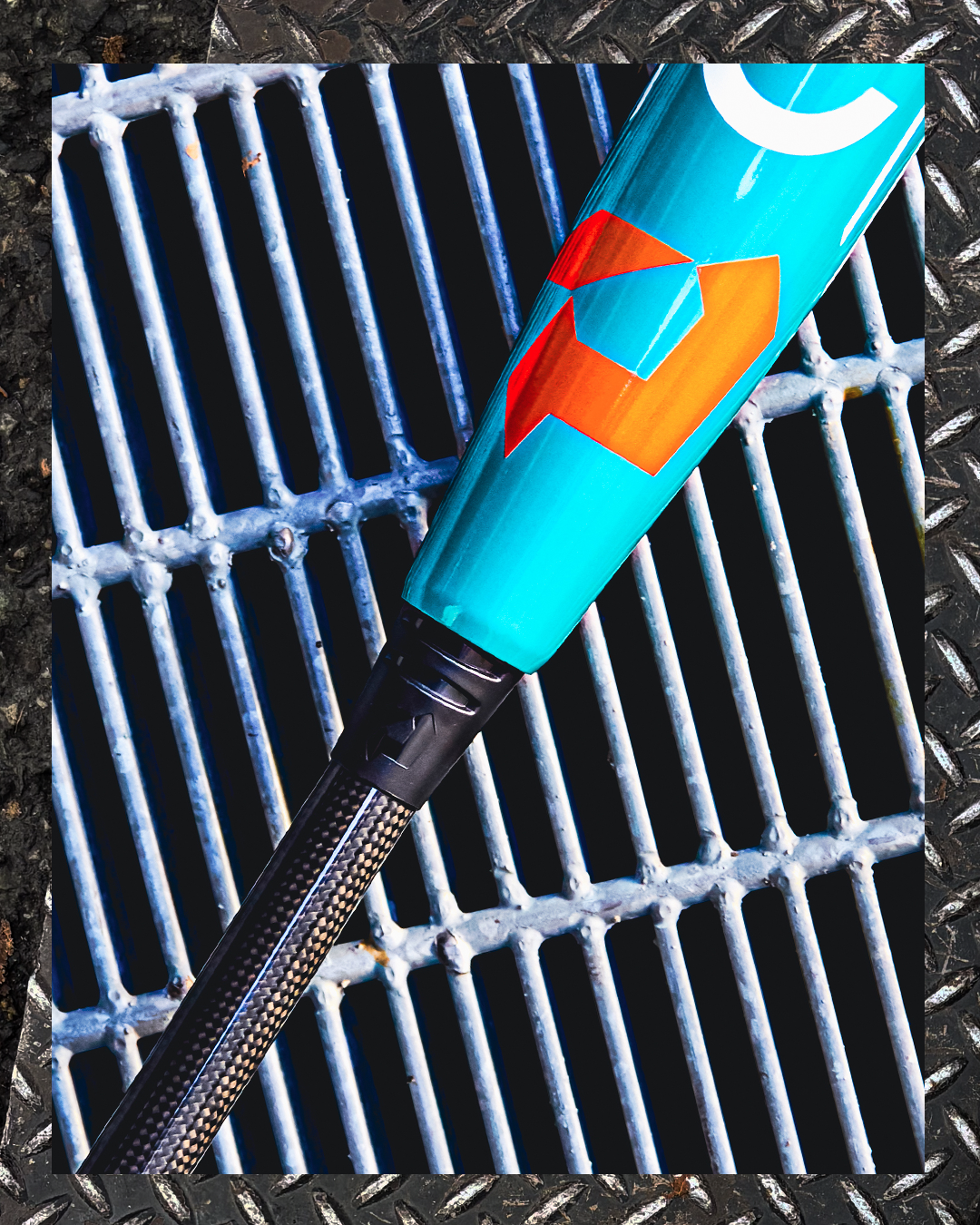 A close-up of the 2026 DeMarini Omega (-3) BBCOR Baseball Bat (WBD2586010) with a turquoise finish and orange geometric design rests on a metal grate bordered by diamond plate.