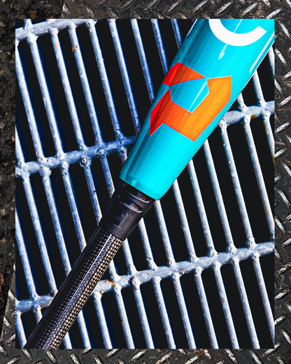 A close-up of the 2026 DeMarini Omega (-3) BBCOR Baseball Bat (WBD2586010) with a turquoise finish and orange geometric design rests on a metal grate bordered by diamond plate.