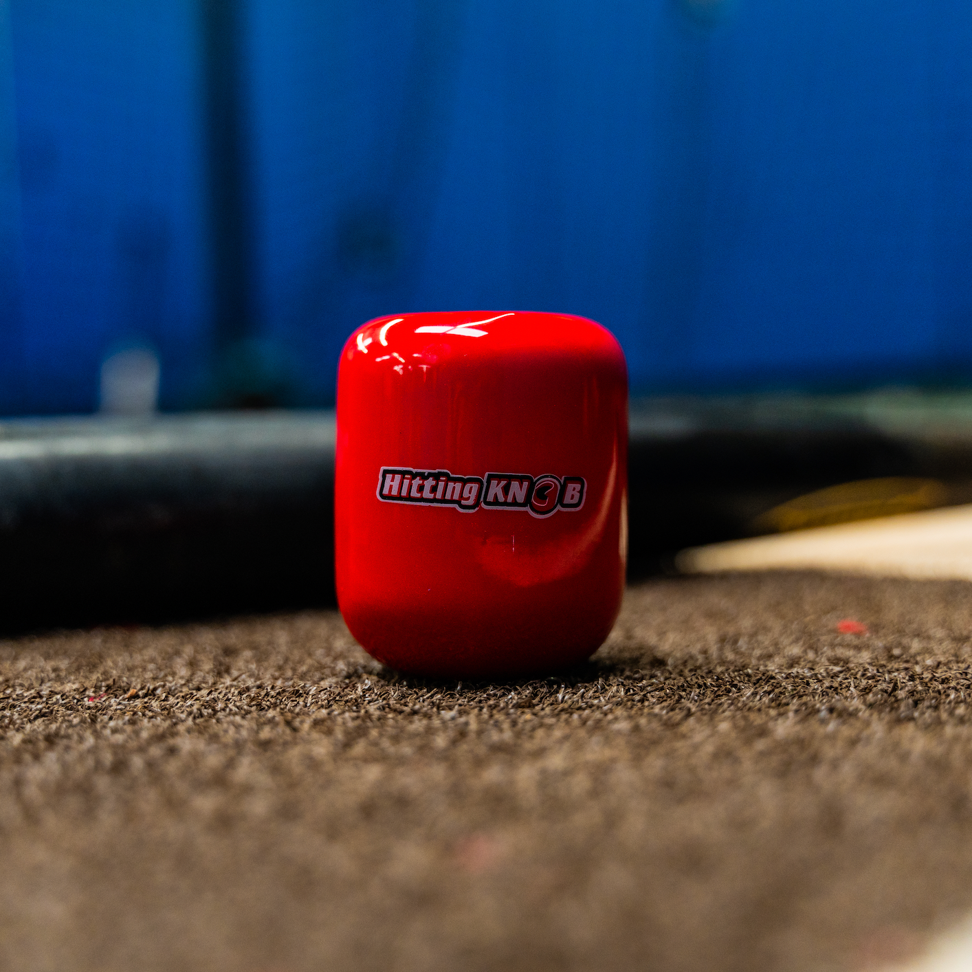 A shiny red The Hitting Knob Bat Weight by Hitting Knob, a baseball training aid that increases bat speed, rests on a textured surface with a blurred blue background.