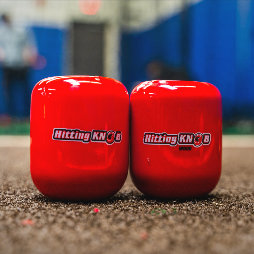 Two bright red, cylindrical Hitting Knob Bat Weights from the Hitting Knob brand rest side by side on a sports field, designed as baseball hitting aids to help players boost bat speed. A blurred indoor background completes the energetic scene.