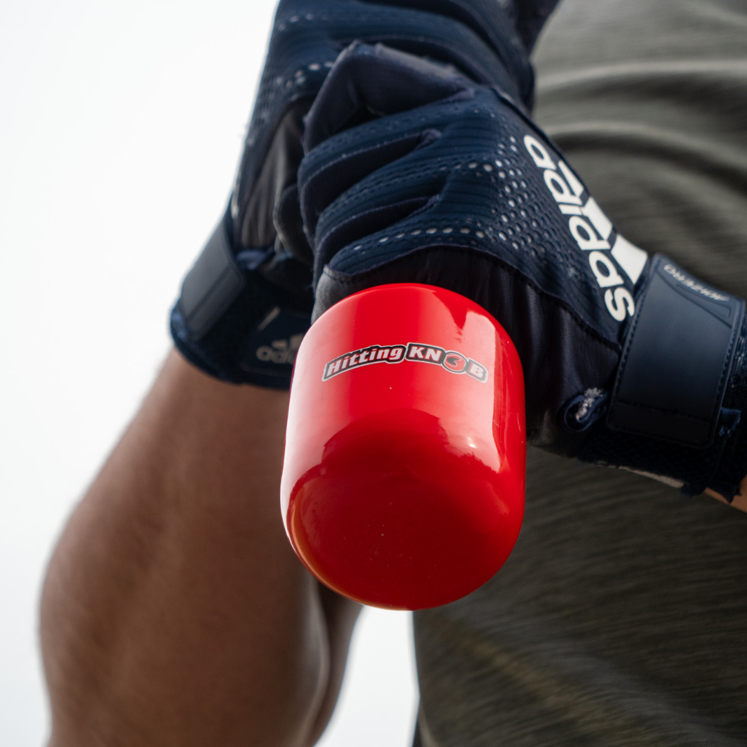 A person wearing black Adidas gloves grips a baseball bat fitted with The Hitting Knob Bat Weight by Hitting Knob, a red training aid designed to help increase bat speed.