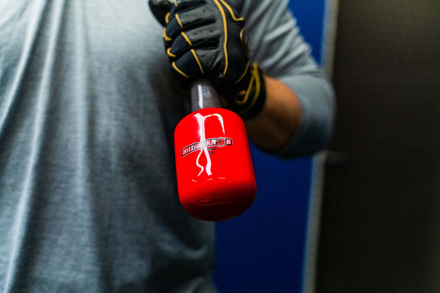 A person in a gray shirt and black gloves holds The Hitting Knob Bat Weight by Hitting Knob—a bright red baseball training aid to increase bat speed—against a blue and black background.