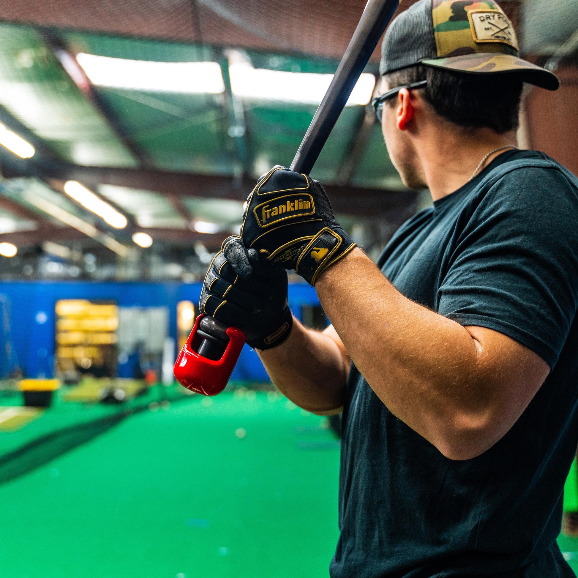 A person in a cap, sunglasses, and batting gloves holds a bat fitted with the Hitting Knob Bat Weight by Hitting Knob in a batting cage with green turf and assorted baseball equipment visible in the background.