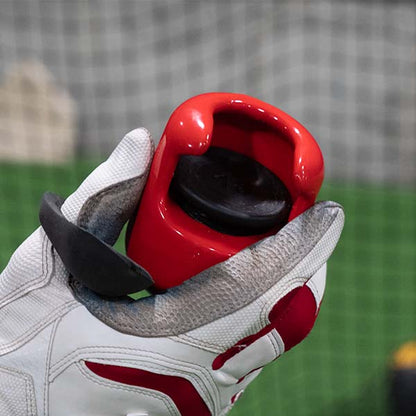 A person wearing white and red sports gloves holds The Hitting Knob Bat Weight by Hitting Knob with a black cap, in front of a blurred sports net and green turf background.