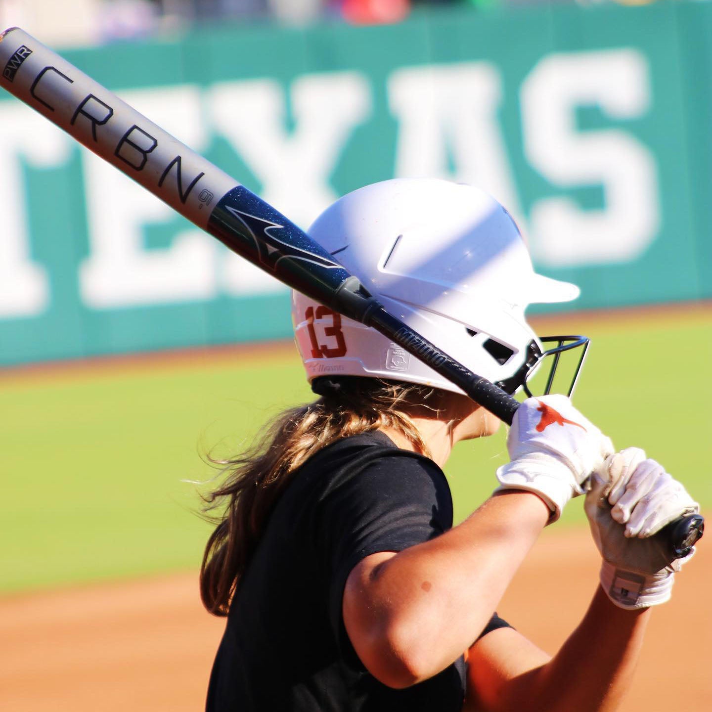 A softball player wearing a white helmet and black jersey with the number 13 prepares to bat, holding a CRBN bat. A blurred green and white sign reading TEXAS is visible in the background.
