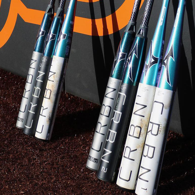 Several Mizuno CRBN baseball bats with teal, black, and silver designs are lined up vertically against a wall on a brown turf surface. An orange graphic is partially visible in the background.
