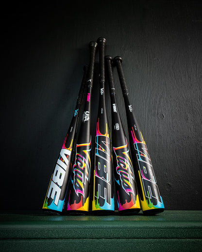 Four 2025 Victus Vibe (-3) BBCOR Baseball Bats by Victus stand propped against a dark wall, each one-piece aluminum bat featuring vibration-reducing knobs, bold stylized text, and vibrant blue, pink, and yellow handle designs.