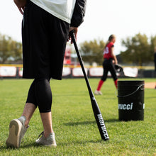 A person stands on grass holding the Marucci Aluminum Fungo 35″ Training Bat: MTRBFA near a Marucci bucket, while a softball player in red and black walks in the background. The bat, made by Marucci, features high-strength alloy construction and an anti-vibration knob for enhanced performance and comfort.