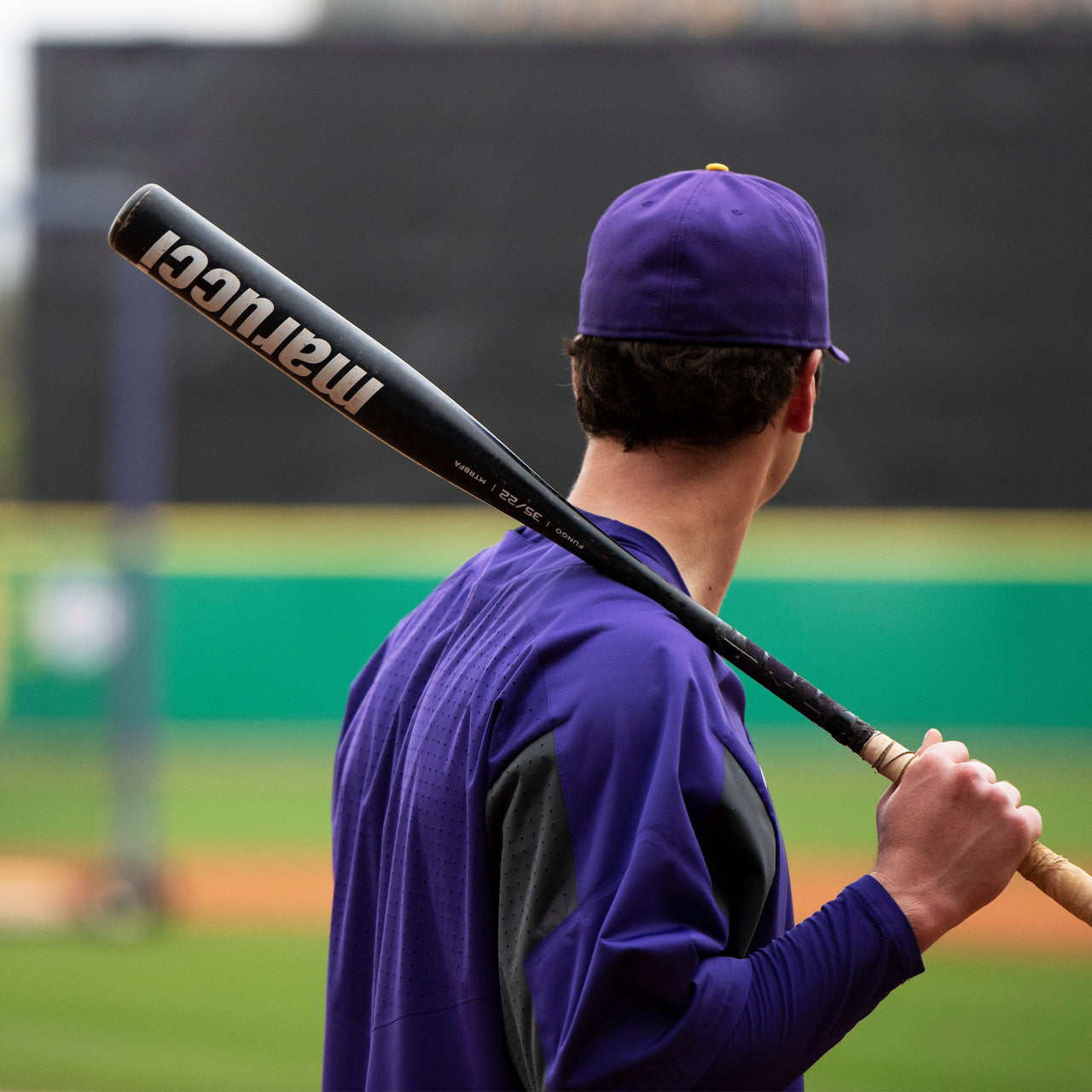 A baseball player in a purple uniform and cap stands on a field, holding a Marucci Aluminum Fungo 35″ Training Bat (MTRBFA) constructed with high-strength alloy over his shoulder, facing away from the camera.