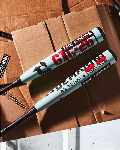 Two light blue DeMarini 2026 The Goods (-3) BBCOR Baseball Bats (WBD2588010) with black handles rest on brown cardboard. These hybrid bats feature red, black, and gray logos and an X14 Alloy Barrel for powerful performance.