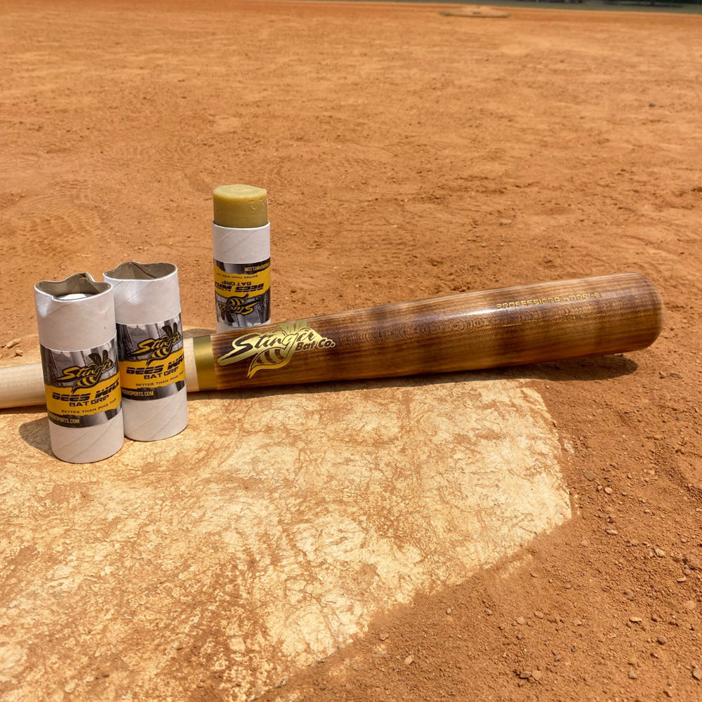 A Stinger Sports BEES WAX Bat Grip and three wrapped grips—offering a natural, pine tar-free hold—are placed on home plate on a sunlit dirt field.
