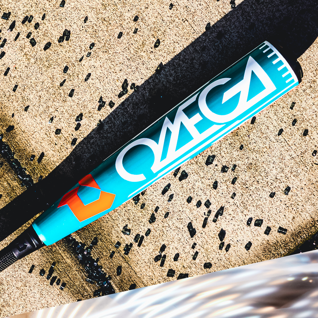A 2026 DeMarini Omega (-3) BBCOR Baseball Bat (WBD2586010) with "OMEGA" in bold white letters rests on concrete, partly in sunlight, while black rubber pellets are scattered around it.