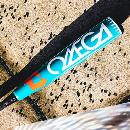 A 2026 DeMarini Omega (-3) BBCOR Baseball Bat (WBD2586010) with "OMEGA" in bold white letters rests on concrete, partly in sunlight, while black rubber pellets are scattered around it.